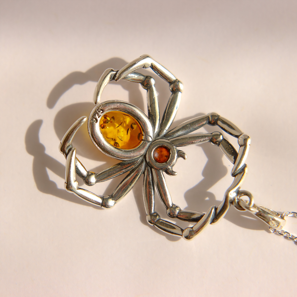 A silver amber spider pendant necklace featuring a large oval amber-yellow gemstone and a small round orange gemstone, displayed on a light beige background with a soft shadow.