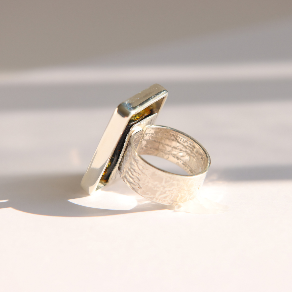 A silver amber ring with a textured band and a geometric, angular flat top, photographed on a white surface with soft shadows.