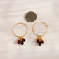 A pair of gold hoop earrings with three dark red teardrop bead drops each, displayed next to a US quarter coin for scale on a light beige background.