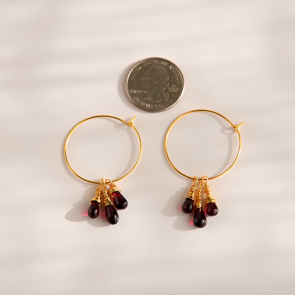 A pair of gold hoop earrings with three dark red teardrop bead drops each, displayed next to a US quarter coin for scale on a light beige background.
