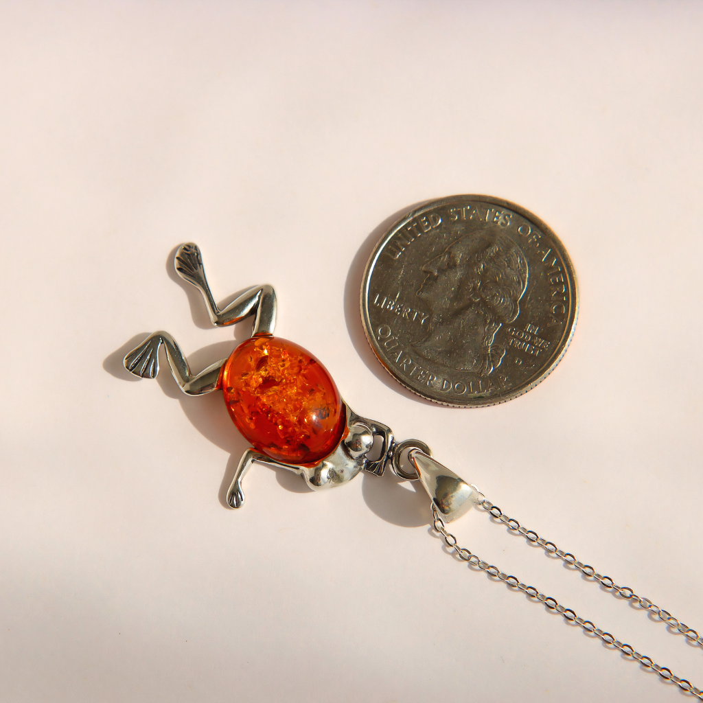A silver amber frog pendant necklace with a polished orange amber center, shown next to a US quarter coin for scale on a light beige background.