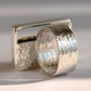 A silver amber ring with a hammered, textured exterior and a smooth, engraved interior, shown next to a matching rectangular silver tag on a light gray background.