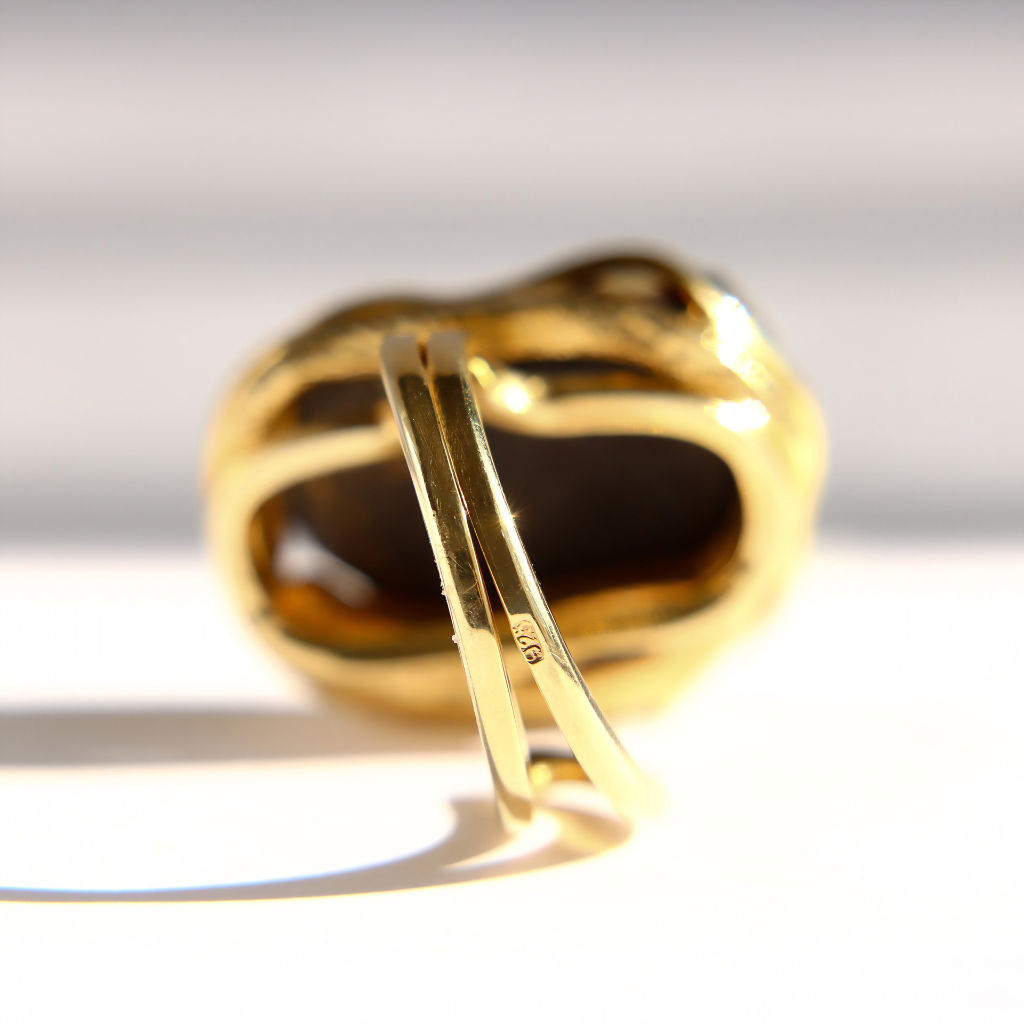 A close-up of a gold amber ring with a large organic-shaped dark black amber stone, photographed on a bright white background with strong sunlight and shadows.