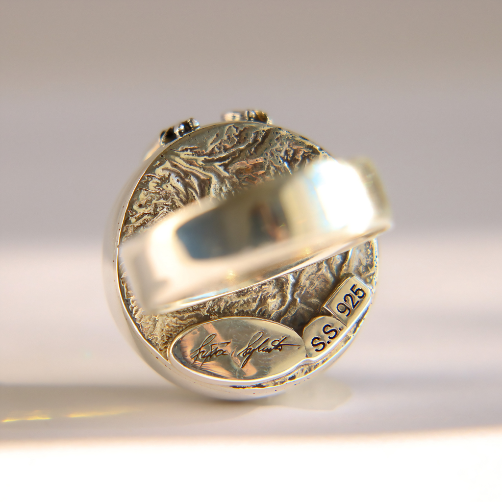 A sterling silver amber ring with an intricate landscape relief design on the band and a polished round center stone, shown on a light gray background.