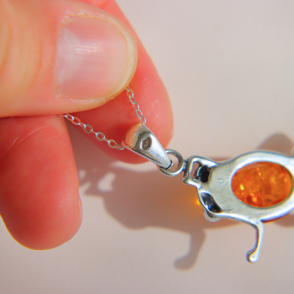 A close-up of a hand holding a silver chain amber frog necklace featuring an orange amber inlay, set against a plain light background.