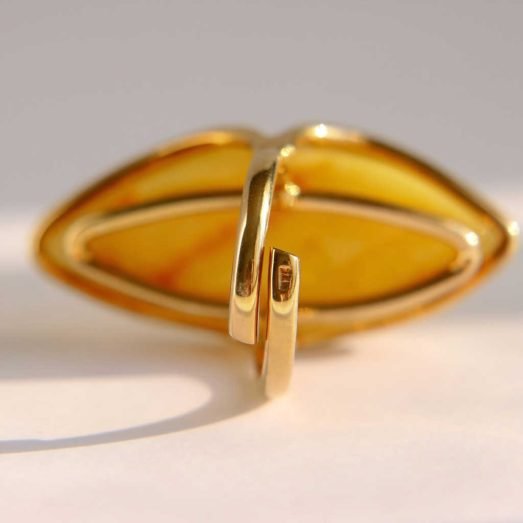 A gold butterscotch amber ring with a large, organic, shield-like openwork design featuring a translucent amber-yellow center, photographed on a light gray background.