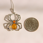 A silver amber spider pendant necklace shown next to a US quarter coin for scale on a light beige background.