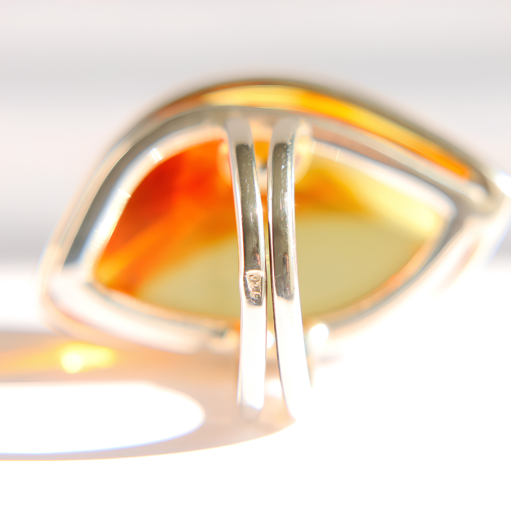 A close-up of a silver amber statement ring with a large, abstract teardrop-shaped amber or honey-colored inlay, with 925 stamp ,photographed against a bright white background.