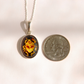 A silver oval pendant amber scarab necklace featuring a yellow and orange beetle design on a dark background, displayed next to a US quarter coin for scale on a light beige surface.