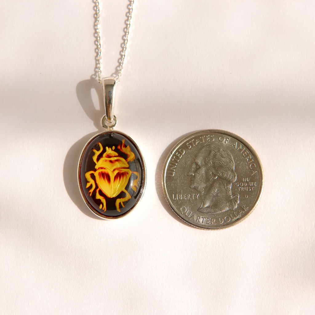 A silver oval pendant amber scarab necklace featuring a yellow and orange beetle design on a dark background, displayed next to a US quarter coin for scale on a light beige surface.