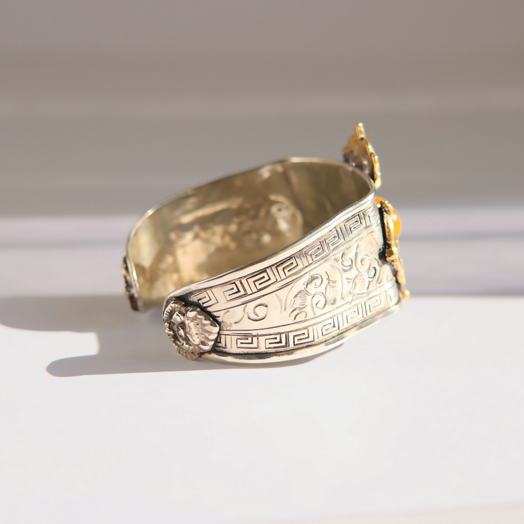 A wide silver amber cuff bracelet with engraved Greek key and scroll motifs, accented by small gold-toned decorative elements on a white background.