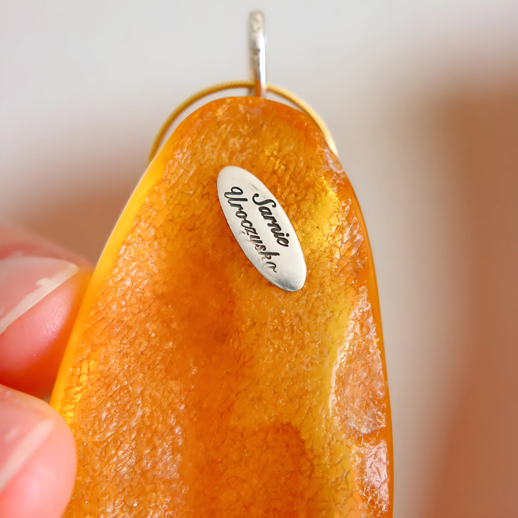 Orange pendant statement necklace with a silver tag held by a hand against a blurred background