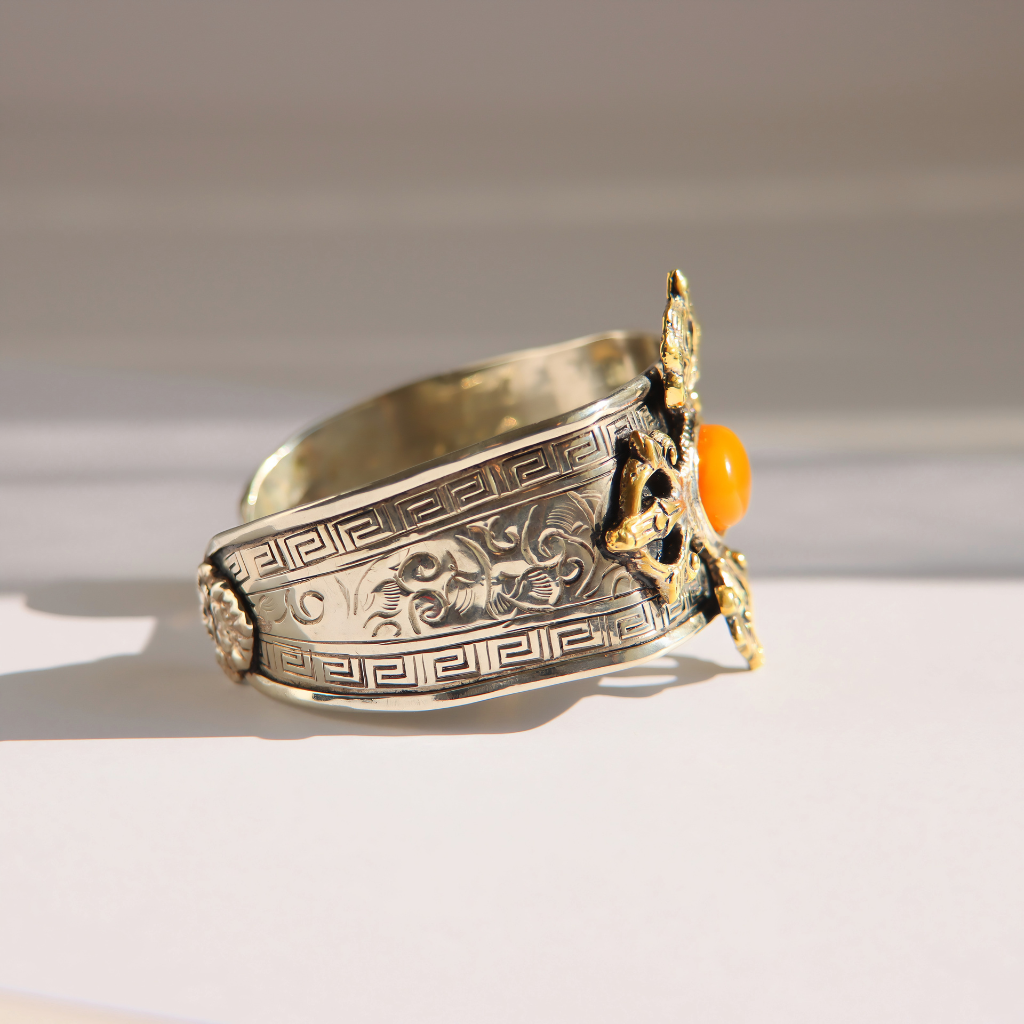 An ornate silver-toned  amber cuff bracelet with engraved floral details, Greek key borders, a gold-toned decorative element, and a round orange bead accent, photographed on a light neutral background.