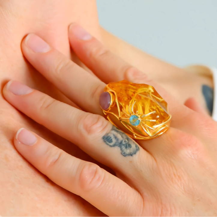 Gold cocktail ring with a blue gemstone on a person's finger against a neutral background