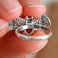 Silver bat ring with red stone held between fingers against a neutral background