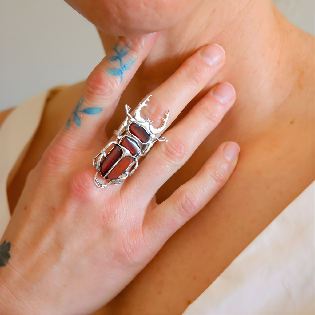 Close-up of a hand wearing a Scarab amber ring with a red stone on a neutral background