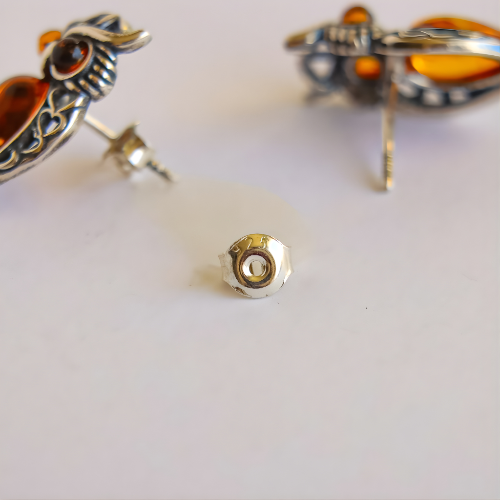 Close-up of silver amber owl earrings with heart designs and a small gold bead on a light background.