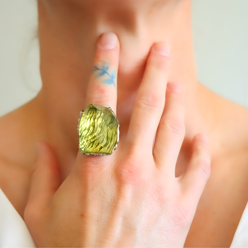 Close-up of a hand wearing a large green amber ring with a blurred background