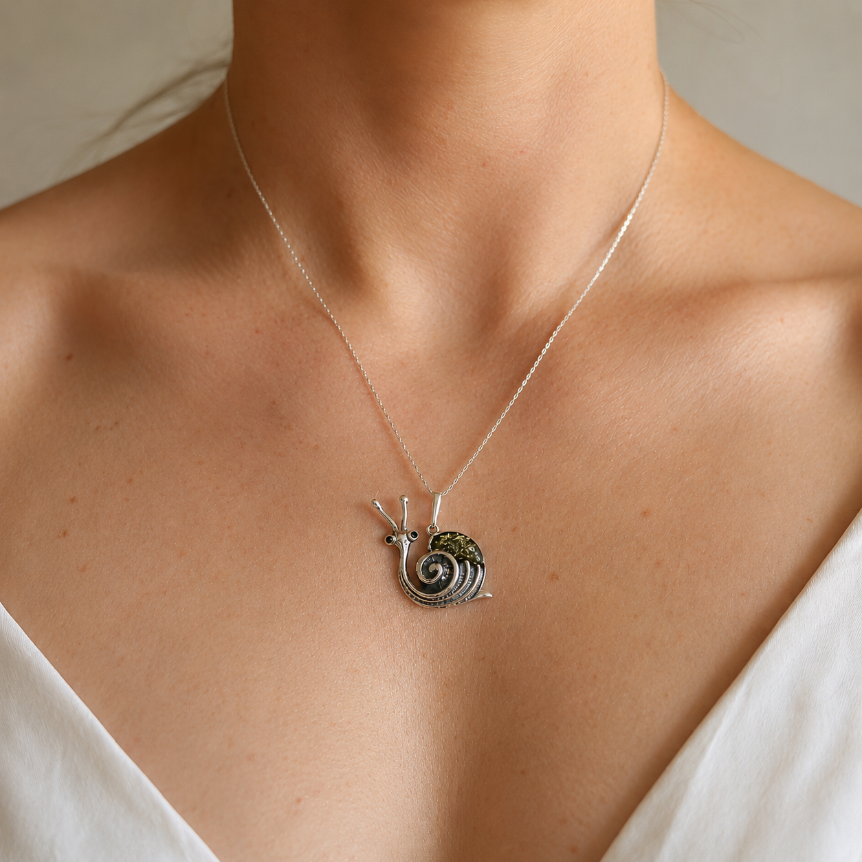 A close-up of a person's neck and upper chest wearing a delicate silver chain necklace with a small amber snail pendant. The pendant has a dark, textured shell with metallic silver details and antennae.