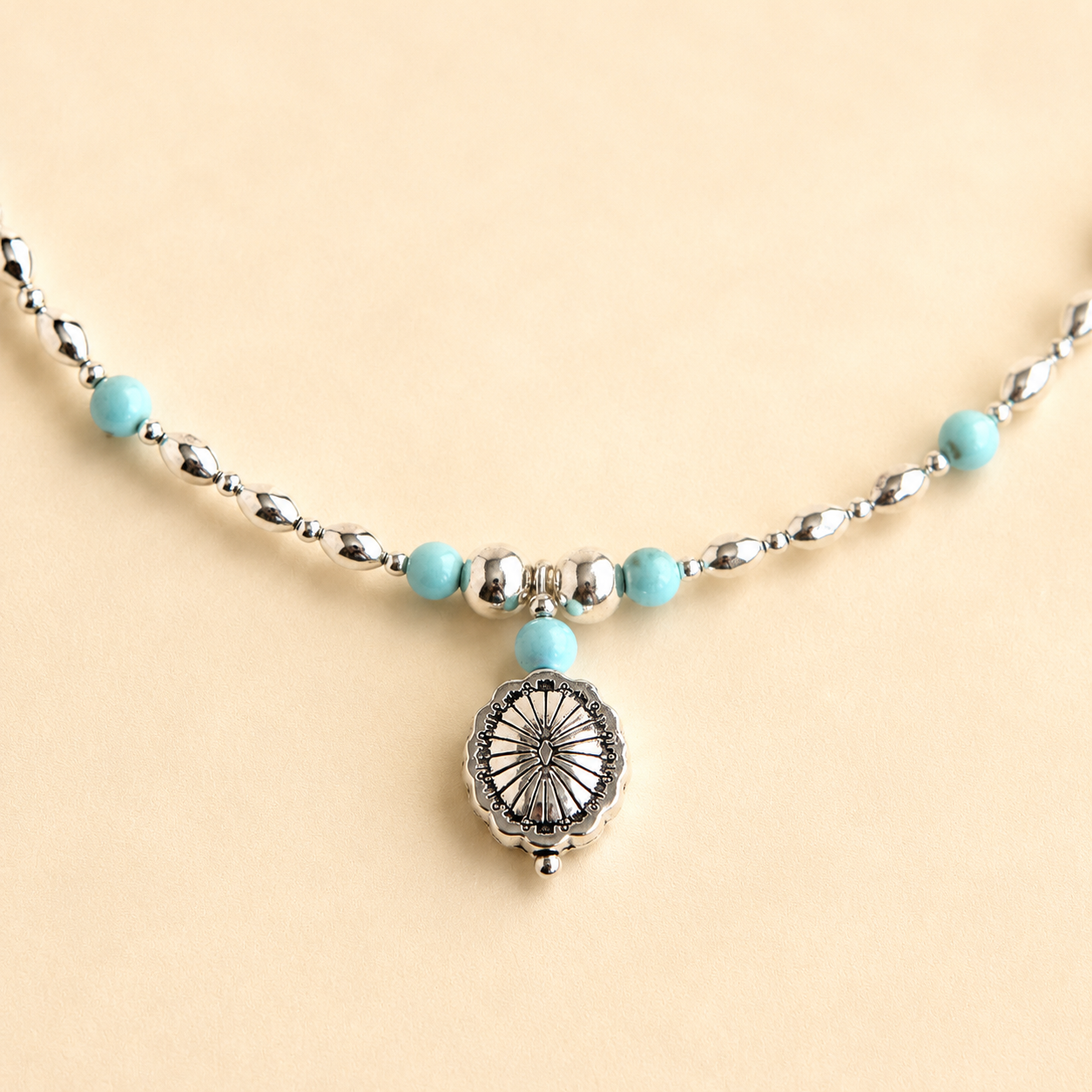 Turquoise necklace with silver beads and silver concho pendant on light beige background
