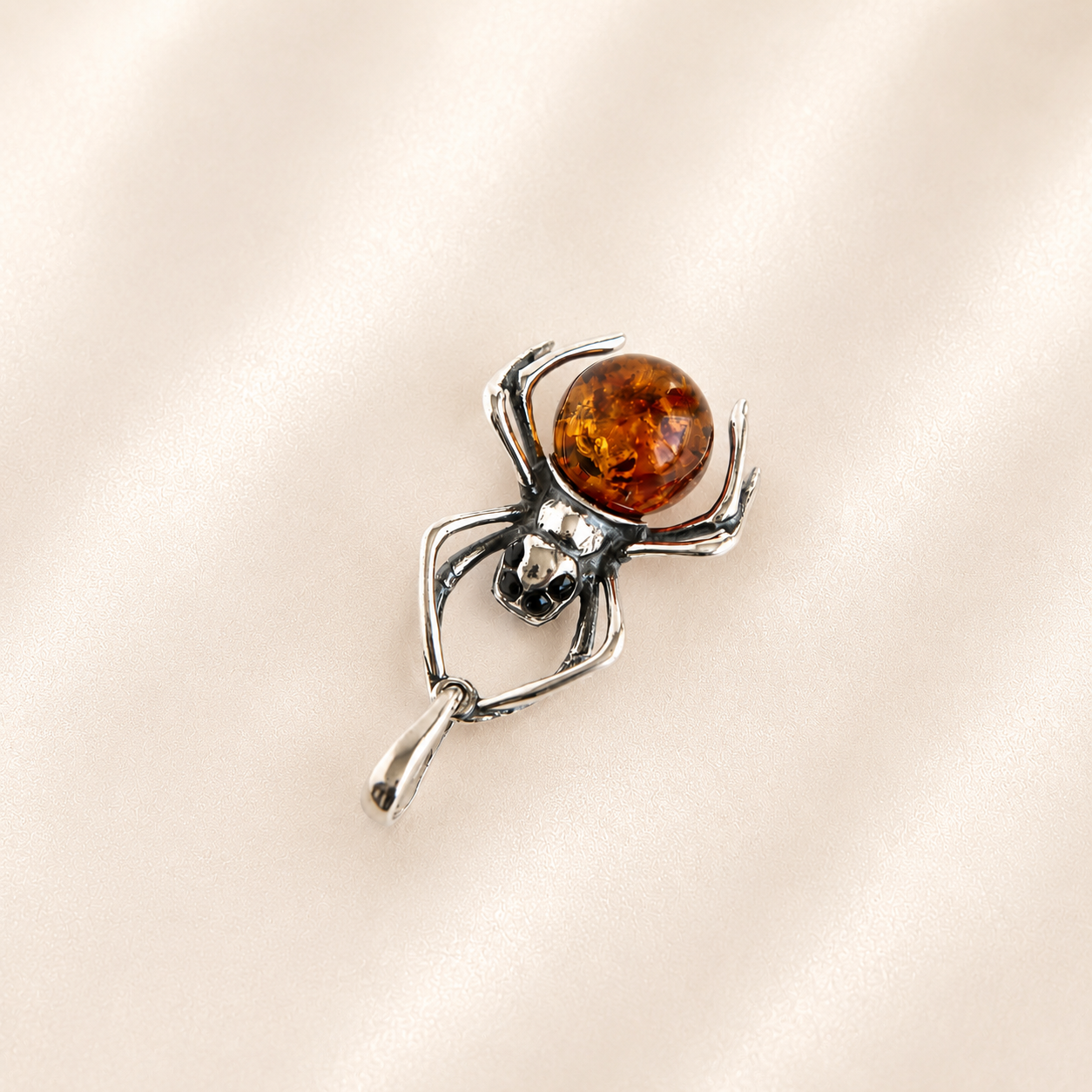 Amber spider pendant with a polished silver-toned metal body and a round amber-orange center, displayed on a soft beige satin fabric background.