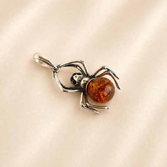 Amber spider pendant with a polished amber-colored orb and silver-toned metal legs, displayed on a soft beige satin fabric background.