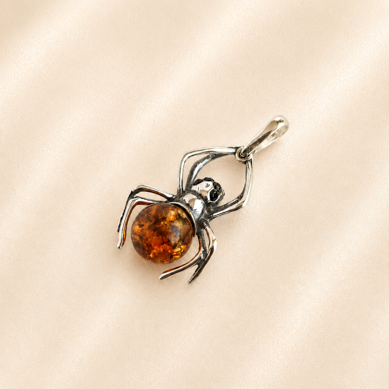 Amber spider pendant with a polished amber-colored round body and silver-toned legs and bail, resting on a soft beige satin fabric background.
