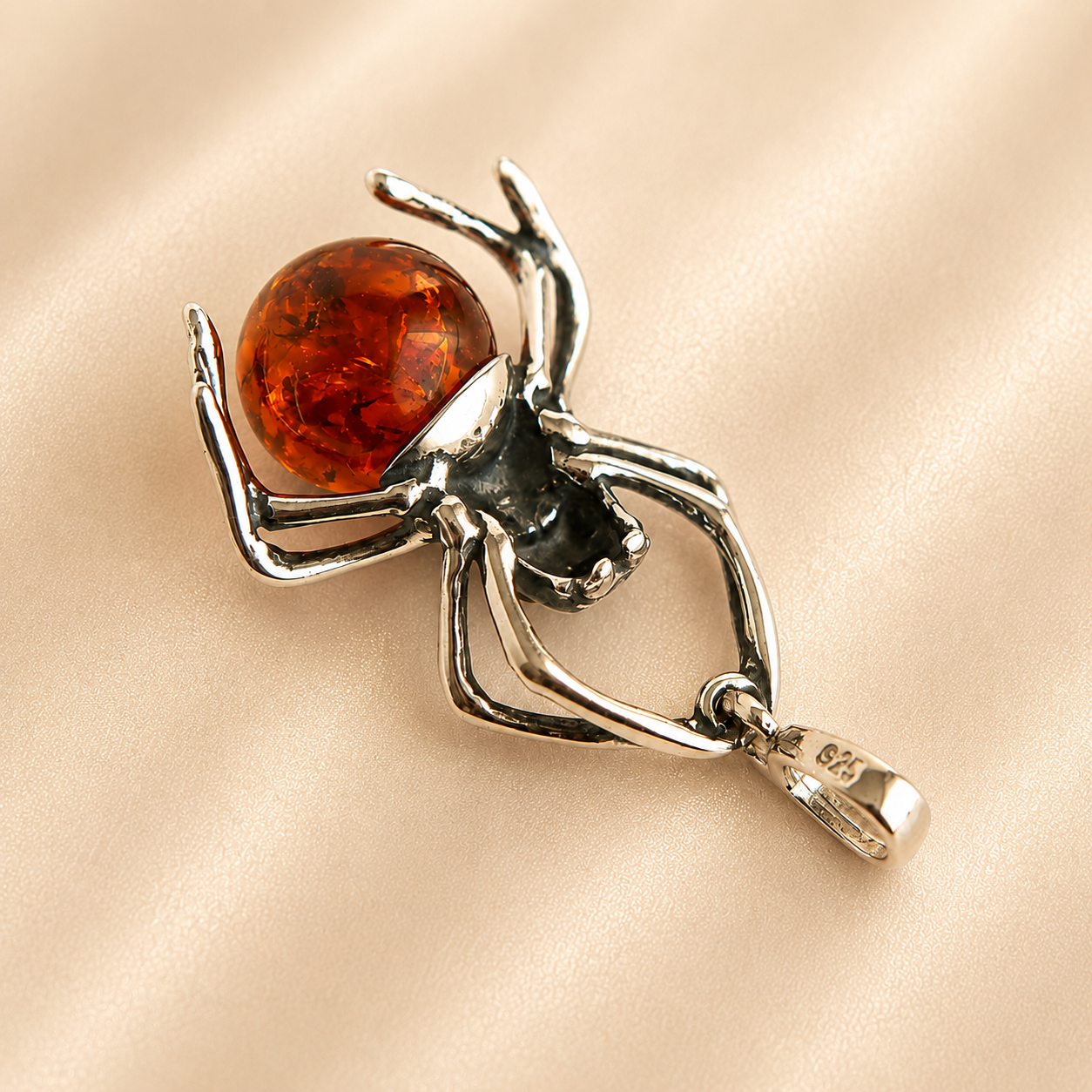 A sterling silver amber spider pendant holding a polished round amber gemstone, displayed on a soft beige satin fabric background.