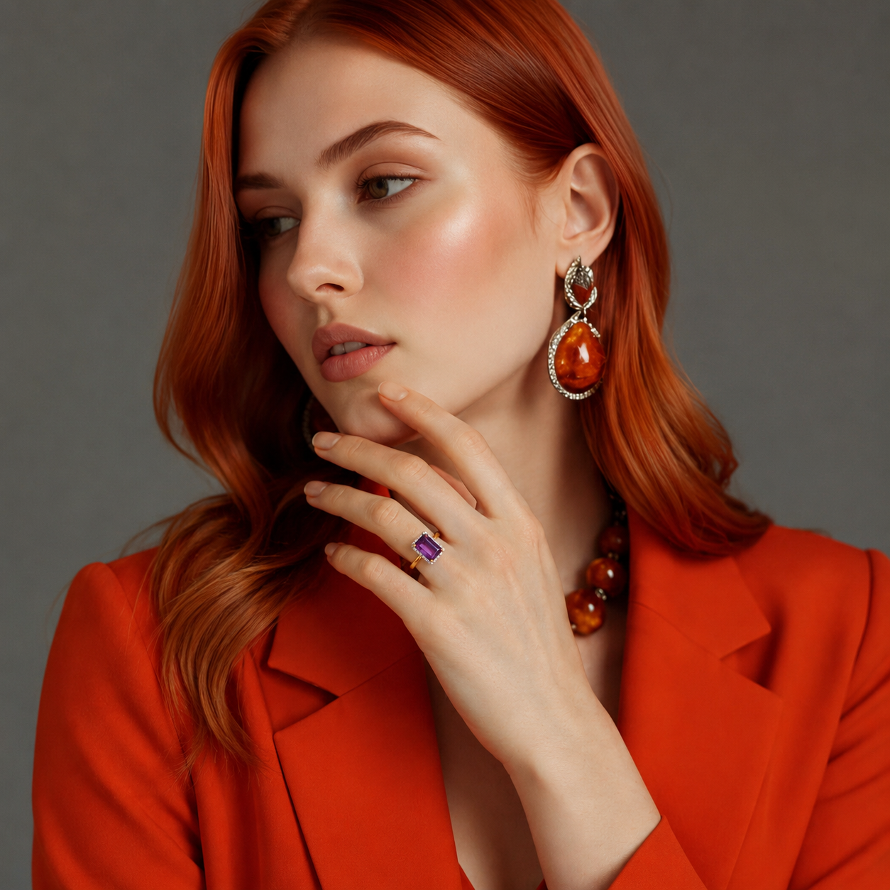Woman wearing a red blazer, amber earrings, and a purple Solitaire ring on a gray background