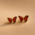 Butterfly gold stud earrings with red centers and gold frames on a beige background