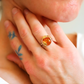 Close-up of a hand wearing a flower ring with an orange gemstone, with a blurred background.