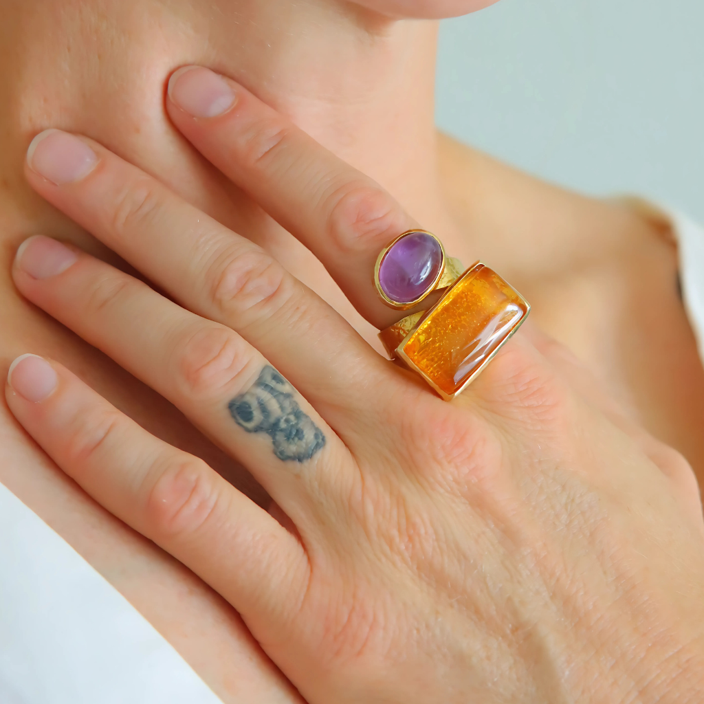 Close-up of a hand wearing a ring with a purple gemstone and amber gemstone on a neutral background