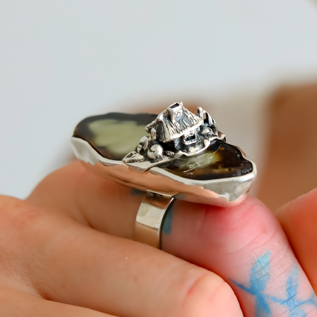 Silver amber ring with a house design on a finger against a neutral background