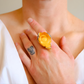 Close-up of a hand wearing a yellow rose ring with a floral design on a neutral background