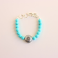 Turquoise beaded howlite bracelet with a silver charm on a light pink background