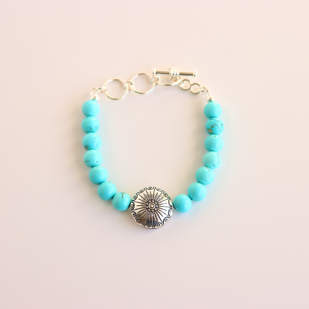 Turquoise beaded howlite bracelet with a silver charm on a light pink background