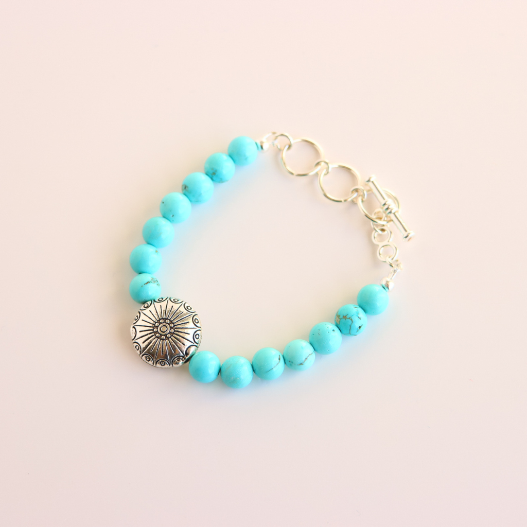 Turquoise beaded howlite bracelet with a silver charm on a light pink background
